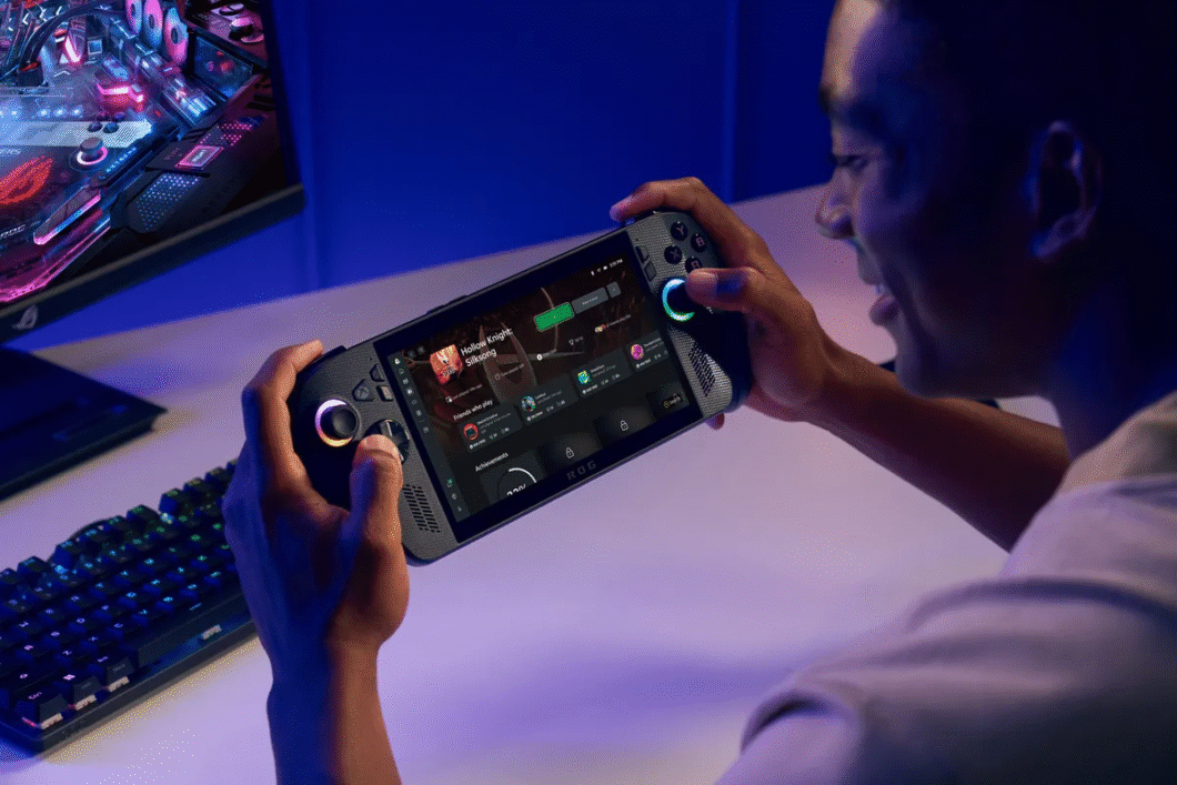 Um homem segura um console Rog Xbox Ally X preto com a tela ligada, exibindo a interface de jogos. O console tem joysticks pretos com anéis coloridos (laranja e verde neon) e botões pretos. Ao fundo, parcialmente visível, há um monitor de computador com uma imagem de um jogo colorido e um teclado. O ambiente é escuro com iluminação em tons de azul e roxo.